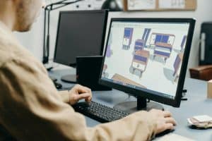 Person working on a computer, viewing a 3D furniture design program, with another monitor and office supplies visible on the desk.