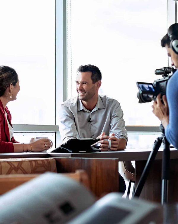 Happy businessman during corporate interview with female journalist. Manager answering question in office. Young woman at work as reporter with business man and cameraman shooting video for broadcast