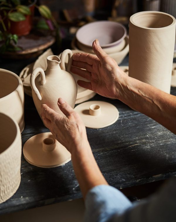Close up of senior woman potter touching white clay pot white sitting at wooden desk with handmade earthenware