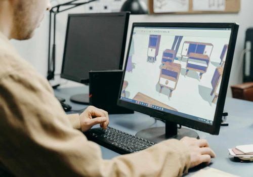 Person working on a computer, viewing a 3D furniture design program, with another monitor and office supplies visible on the desk.