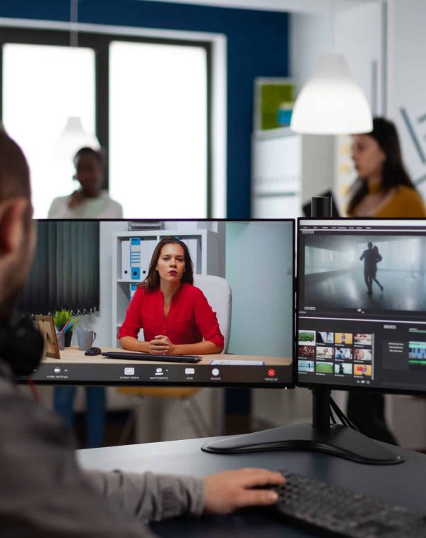 Video editor in web online production conference with project manager on video call editing client work, getting feedback on company movie using post production software on two monitors